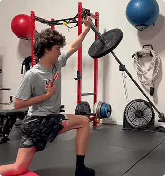 A teenage boy kneeling on his right knee lifting a weight on a pole with his left hand as part of his performance training for athletes program